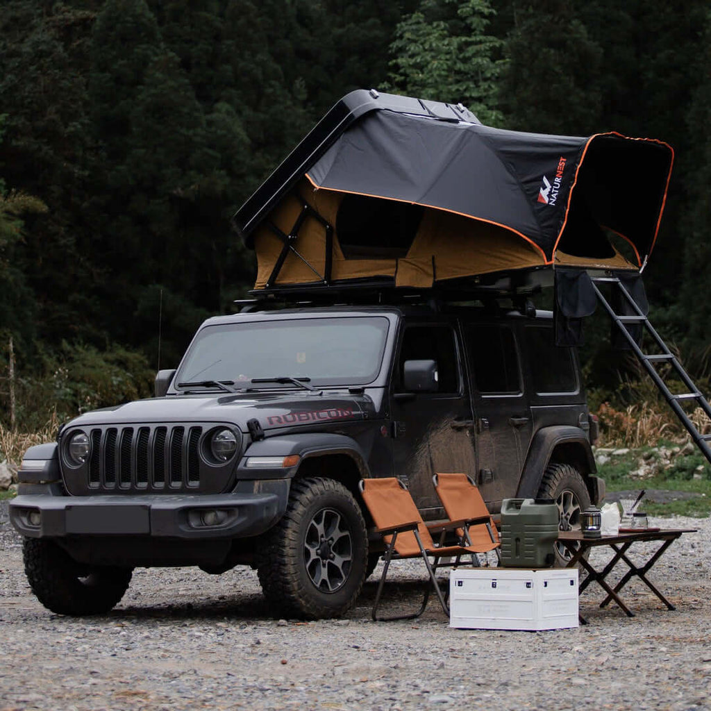 A Black Jeep With A Naturnest Sirius 3 Roof Tent Parked Beside Two Chairs And A Camping Table.