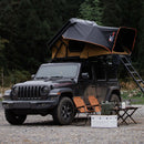 A Black Jeep With A Naturnest Sirius 3 Roof Tent Parked Beside Two Chairs And A Camping Table.