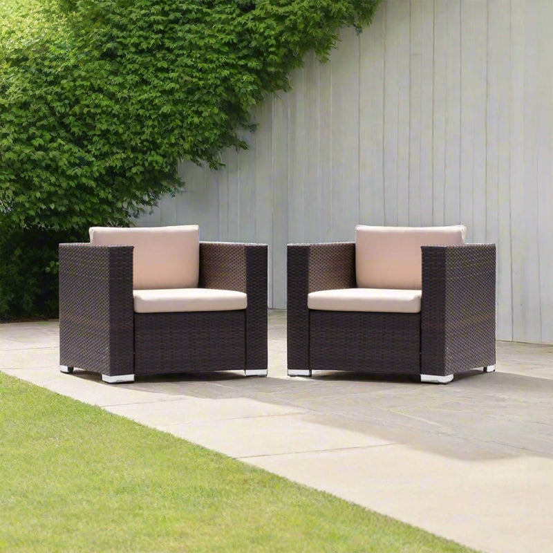 Two Wicker Chairs With Beige Cushions On A Patio Area With Greenery And A Wooden Wall.