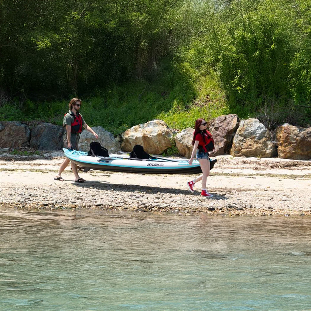 Two People Carrying A Kayak On A Rocky Shoreline With Trees In The Background - Saksby Wholesale