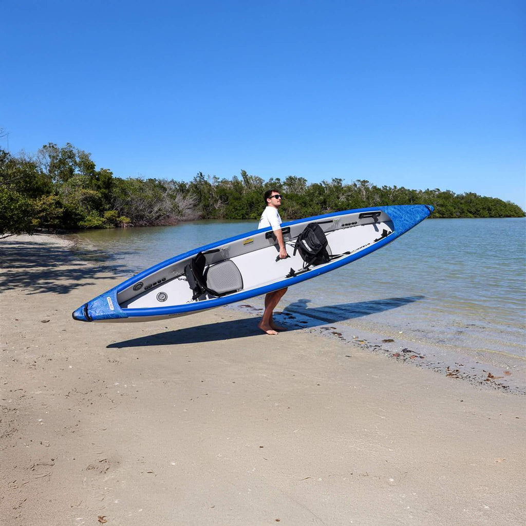 Person Carrying A Blue And White Kayak On A Sandy Beach With Water And Trees In The Background - Saksby Wholesale