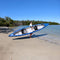 Person Carrying A Blue And White Kayak On A Sandy Beach With Water And Trees In The Background - Saksby Wholesale
