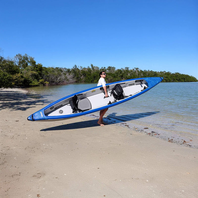 Person Carrying A Blue And White Kayak On A Sandy Beach With Water And Trees In The Background - Saksby Wholesale